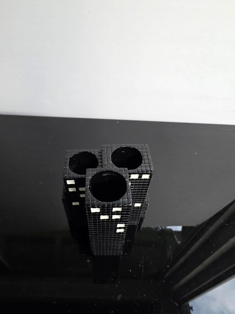 May include: A black ceramic vase with a cityscape design. The vase has three cylindrical openings and is decorated with white rectangular accents to represent windows. The vase is on a reflective black surface.