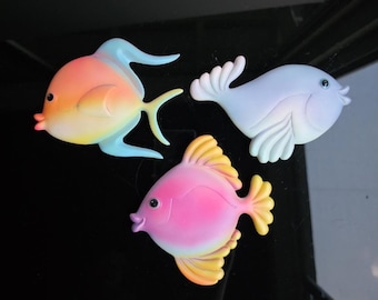 May include: Three colorful fish decorations. One is orange and blue, another is pink and yellow, and the third is light blue. Each fish has a unique design and is set against a black background.