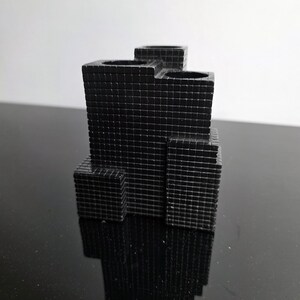 May include: A black, geometric pen holder with a grid-like texture. The pen holder has multiple compartments and a tiered design, resembling a miniature skyscraper. The object is made of a solid material and reflects light.