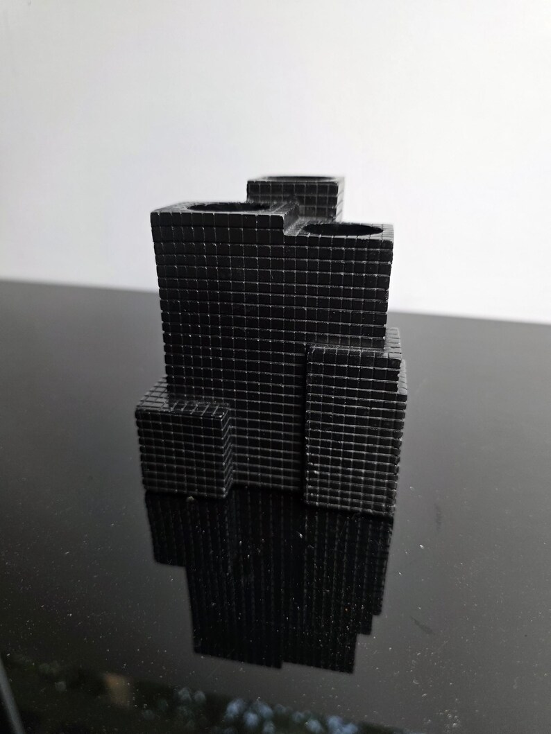 May include: A black, geometric pen holder with a grid-like texture. The pen holder has multiple compartments and a tiered design, resembling a miniature skyscraper. The object is made of a solid material and reflects light.