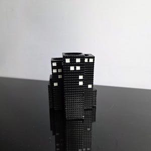 May include: A black, miniature cityscape sculpture with white square windows. The buildings are arranged in a cluster, reflecting on a glossy black surface. The sculpture has a textured surface.