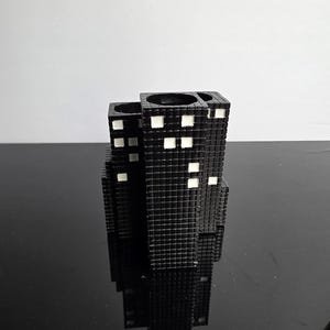May include: Black, cube-shaped toothbrush holder with a grid pattern and white square accents, resembling a cityscape. The holder has multiple openings for toothbrushes and other bathroom essentials. The design is modern and minimalist.