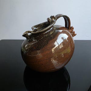 May include: A handcrafted ceramic pitcher with a rounded body and a dark brown glaze. The pitcher features a decorative handle and spout, with a textured surface. The color palette includes shades of brown and gold, creating a rustic aesthetic.