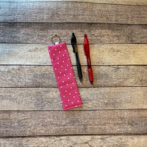 May include: A pink pen holder with white polka dots, a black pen, and a red pen are displayed on a wooden surface. The pen holder has a key ring attached. The pens are black and red.