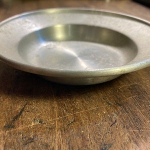 Puede incluir: Un cuenco de metal de color plateado con un borde ligeramente elevado. El cuenco está vacío y tiene una superficie lisa y reflectante.