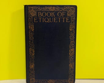 Op de afbeelding: Een vintage boek getiteld "Book of Etiquette" met een donkerblauwe kaft en gouden bloemendetails. De titel staat in gouden letters. Het boek staat rechtop tegen een gele achtergrond.