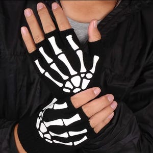 K&ouml;nnte beinhalten: Ein Paar schwarze fingerlose Strickhandschuhe mit einem wei&szlig;en Skeletthand-Aufdruck auf dem Handr&uuml;cken. Das Design zeigt die Knochen der Finger und die Mittelhandknochen als kontrastreiche wei&szlig;e Grafik auf dem schwarzen Stoff der Handschuhe.