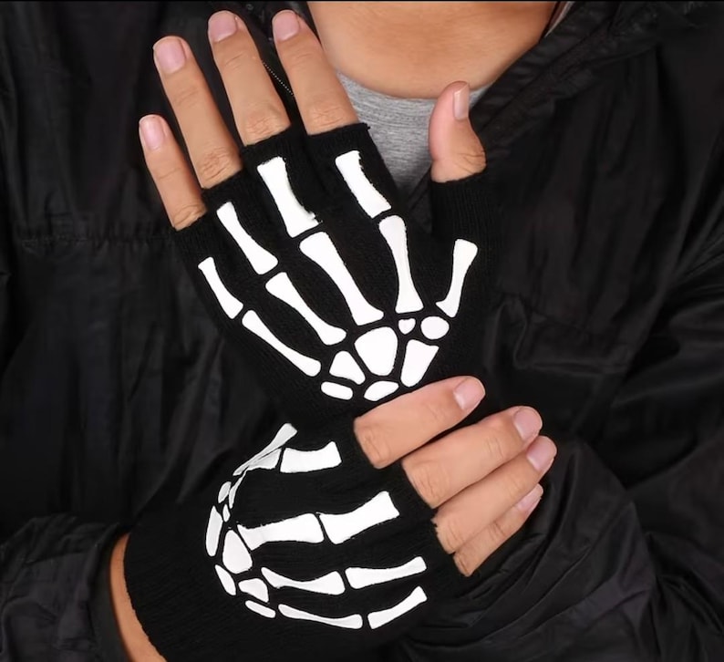 K&ouml;nnte beinhalten: Ein Paar schwarze fingerlose Strickhandschuhe mit einem wei&szlig;en Skeletthand-Aufdruck auf dem Handr&uuml;cken. Das Design zeigt die Knochen der Finger und die Mittelhandknochen als kontrastreiche wei&szlig;e Grafik auf dem schwarzen Stoff der Handschuhe.
