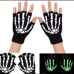 K&ouml;nnte beinhalten: Ein Paar schwarze fingerlose Handschuhe mit einem wei&szlig;en Skeletthand-Knochenaufdruck auf dem Handr&uuml;cken. Die Handschuhe bestehen aus Strickstoff und verf&uuml;gen &uuml;ber eine Leuchtfunktion, die das Knochenmuster im Dunkeln hellgr&uuml;n erstrahlen l&auml;sst.