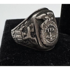 May include: A silver-toned metal ring featuring an embossed emblem of an eagle perched atop a shield with the letters FFA. The design includes the text Vocational Agriculture and FFA, with an owl and a plow depicted within the central circular crest.