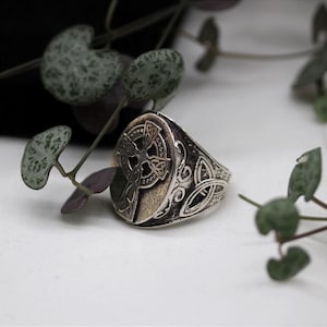 May include: A silver-toned metal ring featuring a circular face with an embossed Celtic cross design. The band of the ring displays intricate, raised knotwork patterns along the sides, with a polished finish and darkened recessed areas.