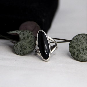 May include: A silver ring featuring a smooth, elongated oval black stone set vertically in the center. The band of the ring splits into two separate silver strands on each side, creating an open, layered design that frames the central stone.