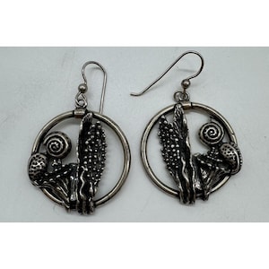 May include: A pair of silver earrings. Each earring has a circular frame with botanical designs, including a spiral, textured pods, and a central, spiky element. The earrings have a hook closure.