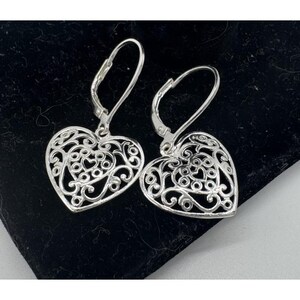 May include: A pair of silver-toned heart-shaped earrings. Each earring features an open filigree design with intricate swirls and heart shapes. The earrings have lever-back closures and are displayed on a black surface.