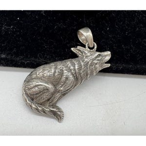 Può includere: Un ciondolo d'argento a forma di lupo che ulula. Il ciondolo è dettagliato con una texture di pelliccia e un anello per una catena. Il lupo è raffigurato seduto, con la testa alzata e la bocca aperta.