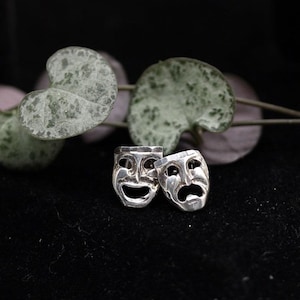 May include: A pair of silver stud earrings featuring the comedy and tragedy theater masks. Each mask is crafted with detailed facial expressions, including a smiling mouth on one and a downturned, sad mouth on the other, finished in a polished metallic silver tone.