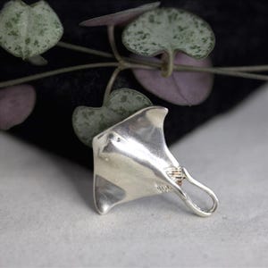 Può includere: Un ciondolo in argento lucido a forma di manta con una superficie liscia e riflettente e una coda curva che forma un occhiello per l'aggancio. Il design presenta un corpo largo e piatto con ali appuntite e una trama dettagliata vicino alla coda.