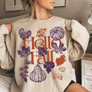 Peut inclure: Sweat-shirt beige avec un motif sur le thème de l'automne. Le graphisme comprend le texte "Hello Fall" en orange, ainsi que des illustrations de citrouilles et de feuilles en violet et orange. Le sweat-shirt est de style col rond.