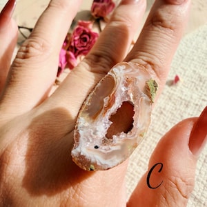 May include: A ring featuring a large, irregular, polished agate slice with a central geode cavity. The stone displays translucent bands of white, tan, and brown, with a small letter C printed in black on the lower right side of the setting.