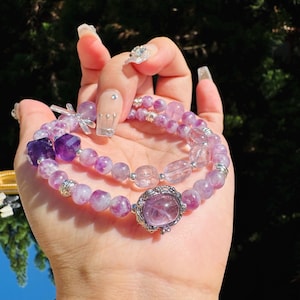 May include: Two amethyst gemstone bracelets with silver accents. The bracelets are layered on top of each other and feature a variety of purple and clear beads.