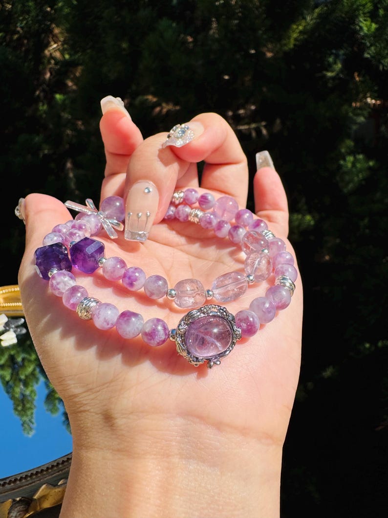 May include: Two amethyst gemstone bracelets with silver accents. The bracelets are layered on top of each other and feature a variety of purple and clear beads.