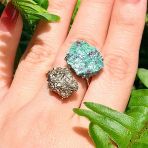 May include: Two gemstone rings. One ring features a rough, teal-colored stone with red inclusions. The other ring has a silver-colored, textured stone. Both rings are set on silver bands. The background includes green foliage.