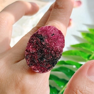 May include: A raw, oval-shaped ring featuring a deep burgundy and black stone. The stone has a textured surface and is set on a simple band. The ring is a unique piece of jewelry.