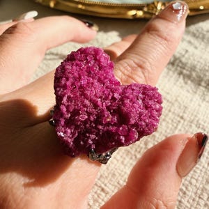 May include: A unique ring featuring a heart-shaped, textured, magenta-colored stone. The stone is mounted on a silver-colored band with decorative accents. The ring is shown on a person's finger, with a soft, neutral background.