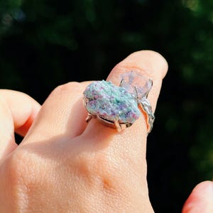 May include: A silver ring featuring a large, textured gemstone with shades of blue, pink, and green. A clear, faceted stone sits beside the larger stone. The ring is worn on a finger, with a blurred green background.