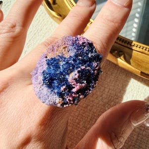 May include: A large, statement ring featuring a cluster of colorful crystals. The ring's focal point is a textured, oval-shaped stone with shades of blue, purple, pink, and gold. The ring is set on a silver band, creating a unique and eye-catching accessory.