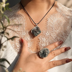 May include: A necklace featuring a raw, dark green druzy crystal pendant on a thin black cord, paired with a matching raw druzy crystal ring. The jewellery set is worn over a white lace-trimmed garment, showcasing the textured, crystalline surfaces.
