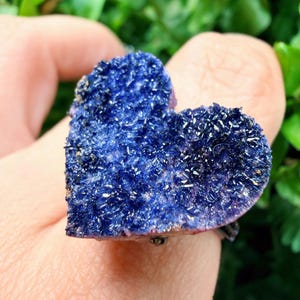 May include: A heart-shaped, deep blue crystal ring. The crystal has a textured surface with small, sparkling inclusions. The ring is held up by a person's finger, with green foliage in the background.