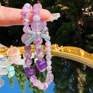 May include: Two purple and pink gemstone bracelets with a silver bow charm. The bracelets are made of round and square beads.
