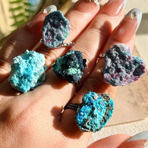 May include: Five rings featuring raw, unpolished mineral stones in various colors including bright turquoise, deep black with green flecks, and mottled pink and blue. Each stone is set on a simple metal band, showcasing natural, jagged textures.