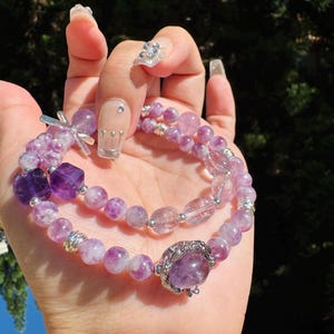May include: Two amethyst bracelets with silver accents. The top bracelet has a silver charm with a cross design. The bottom bracelet has a silver charm with a large amethyst stone.