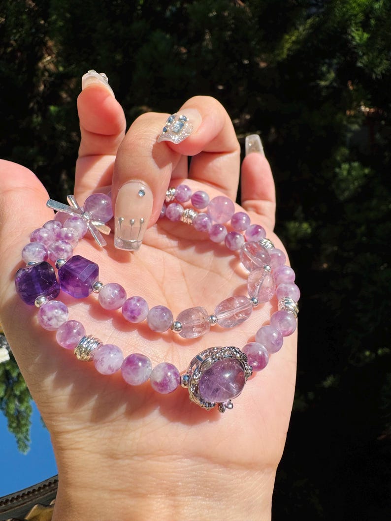 May include: Two amethyst bracelets with silver accents. The top bracelet has a silver charm with a cross design. The bottom bracelet has a silver charm with a large amethyst stone.