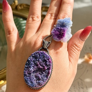 May include: A large oval pendant featuring a deep purple druzy stone set in a silver-toned frame with a panther head bail, paired with a matching ring showcasing a smaller, irregular blue and purple druzy crystal cluster.