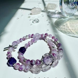 May include: Two amethyst crystal bracelets with silver accents. One bracelet has a cross charm and the other has a round charm with a floral design. The bracelets are on a white surface with other crystals.