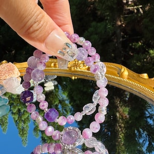 May include: Two purple and clear crystal bracelets with silver accents. The bracelets are made of amethyst and clear quartz beads.