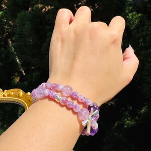 May include: Two layered bracelets with purple and white gemstone beads. The top bracelet has a silver cross charm.