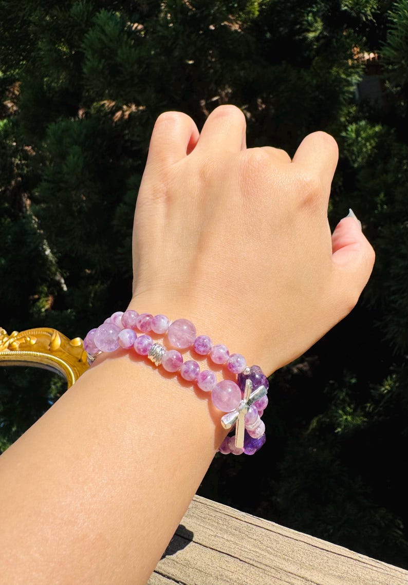 May include: Two layered bracelets with purple and white gemstone beads. The top bracelet has a silver cross charm.