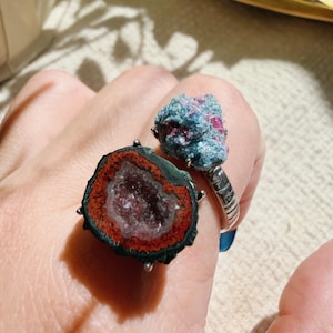 May include: Two gemstone rings. One ring features a geode slice with red, orange, and white colours. The other ring has a cluster of blue and pink crystals. Both rings have silver bands.