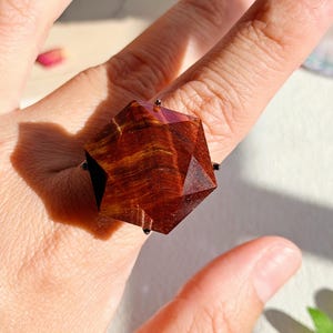 May include: A ring featuring a large, geometric, faceted gemstone with a deep reddish-brown color and natural wood-like grain patterns. The stone is set in a dark metal frame with small prongs securing the multifaceted, star-shaped structure.
