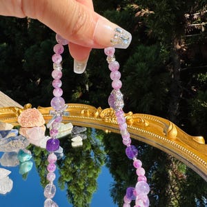 May include: A purple and clear crystal beaded necklace with silver accents. The necklace is made of multiple strands of beads and has a charm in the center.