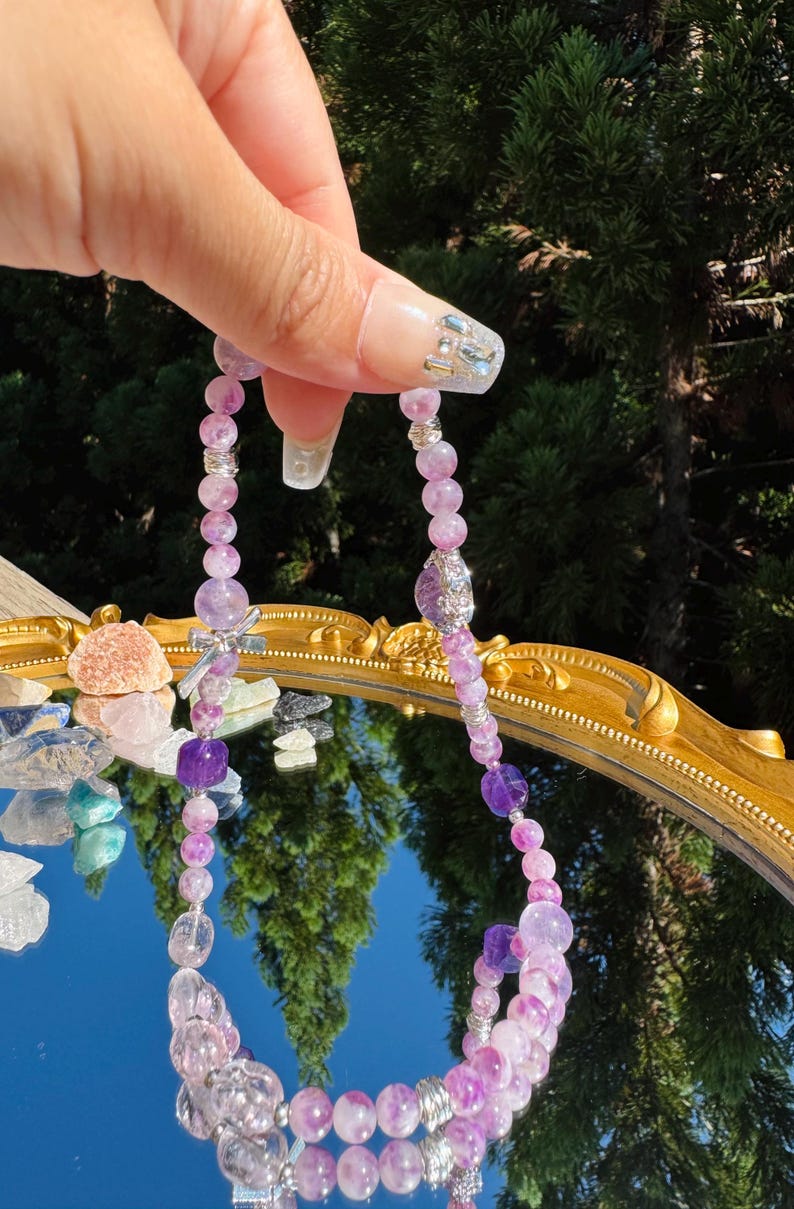May include: A purple and clear crystal beaded necklace with silver accents. The necklace is made of multiple strands of beads and has a charm in the center.