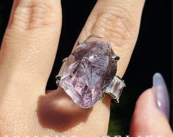 Puede incluir: Un anillo con un gran cristal de amatista morado en bruto montado en una garra de metal plateado, flanqueado por dos piedras más pequeñas, claras y de corte rectangular. El texto JOMONO ATELIER es visible en la parte inferior.