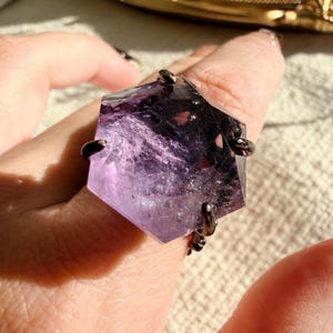 May include: A large, faceted amethyst ring. The purple gemstone is held in place by a dark metal setting. The ring has a geometric shape and is set against a neutral background.