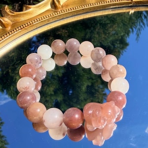 Könnte beinhalten: Ein Armband mit Perlen in Erdtönen, darunter weiße, rosa und braune Perlen. Es hat einen hellorangefarbenen, geschnitzten Bärenanhänger. Das Armband wird auf einer reflektierenden Oberfläche mit einem goldenen, verzierten Rahmen präsentiert.