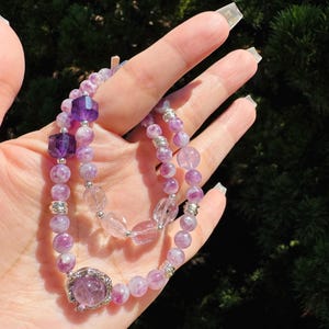 May include: Two purple and white gemstone bracelets with silver accents. The bracelets are made of small, round beads and feature a larger, faceted gemstone in the center.