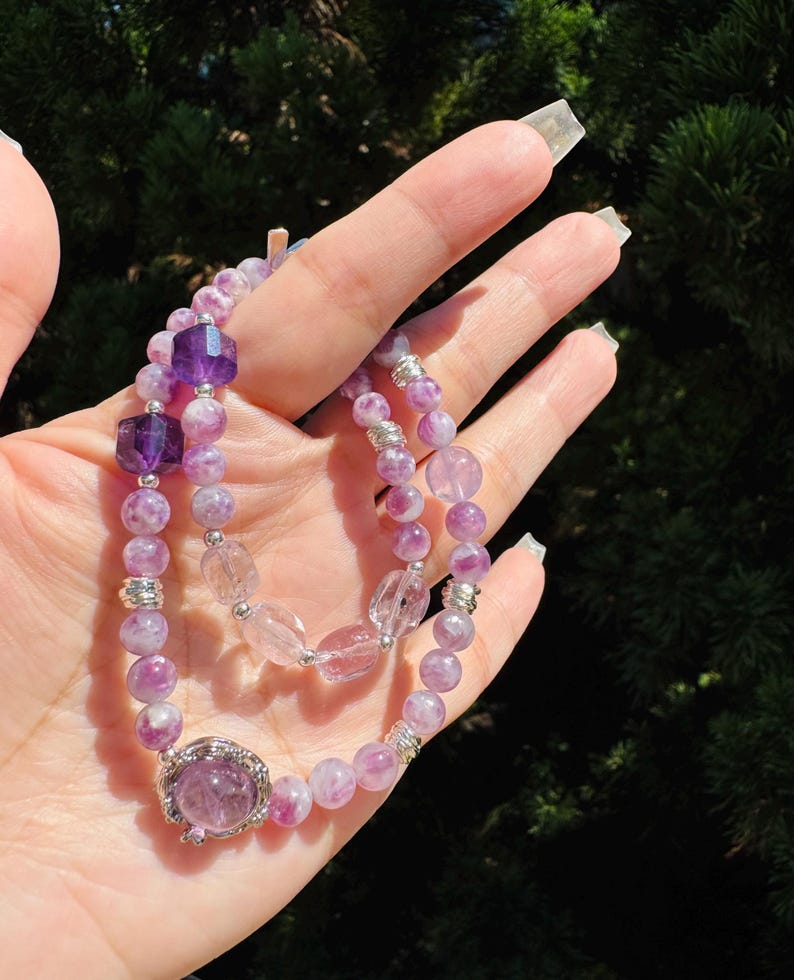 May include: Two purple and white gemstone bracelets with silver accents. The bracelets are made of small, round beads and feature a larger, faceted gemstone in the center.